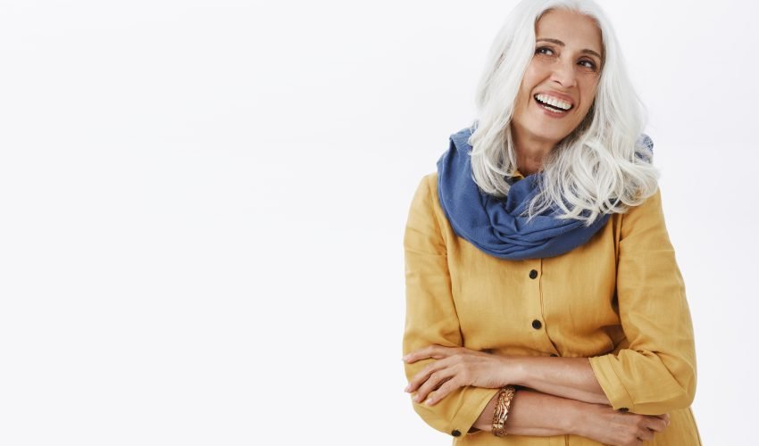 Waist-up shot of charming delighted attractive and feminine elderly female with white hair laughing joyfully wearing stylish blue scarf over neck and yellow trench coat gazing left amused and pleased