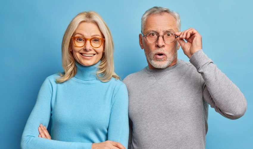 Cheerful middle aged woman and her husband pensioner reacts on shocking news keeps hand on spectacles isolated over blue background. Family age and emotions concept. Senior couple at pose home