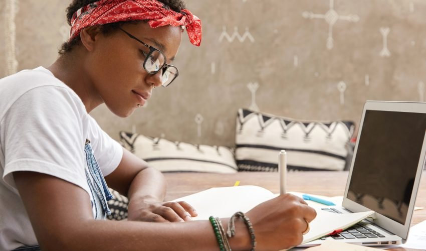 Intelligent ethnic female person with dark skin, concentrated look, records information on paper, sits in front of opened laptop with blank screen for your advertising content, makes research alone