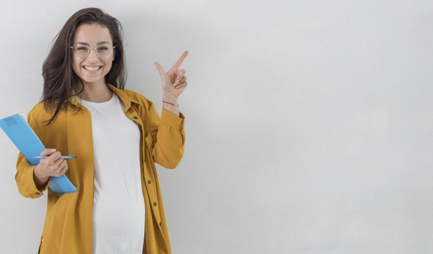 smiley-pregnant-woman-holding-clipboard-pointing-up-with-copy-space