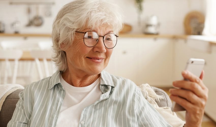Happy joyful female pensioner in round eyeglasses surfing internet on cell phone, looking at mobile’s screen with broad smile, booking plane tickets, planning trip or scrolling pics via social network