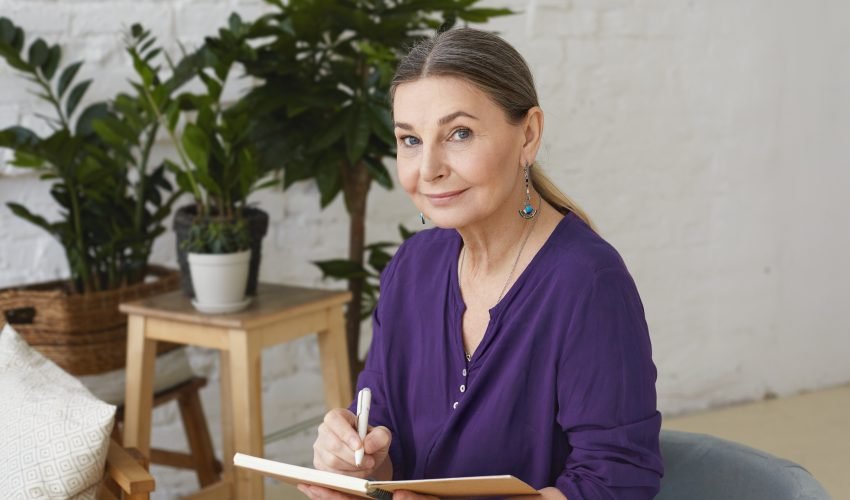 Portrait of busy modern 50 yeard old middle aged woman in violet shirt writing in copybook, making plans, looking at camera with positive friendly smile, sitting on chair, surrounded with green plants