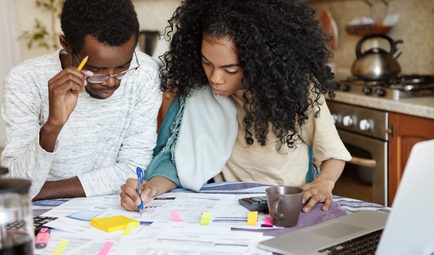 Young confident African housewife with Afro hairstyle helping her husband to manage domestic finances, calculating and making notes with pen, both sitting at kitchen table with laptop and papers