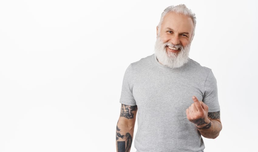 Come closer. Happy handsome old man with tattoos beckon to approach, flick finger, smiling and laughing while luring people, inviting go with him, standing over white background