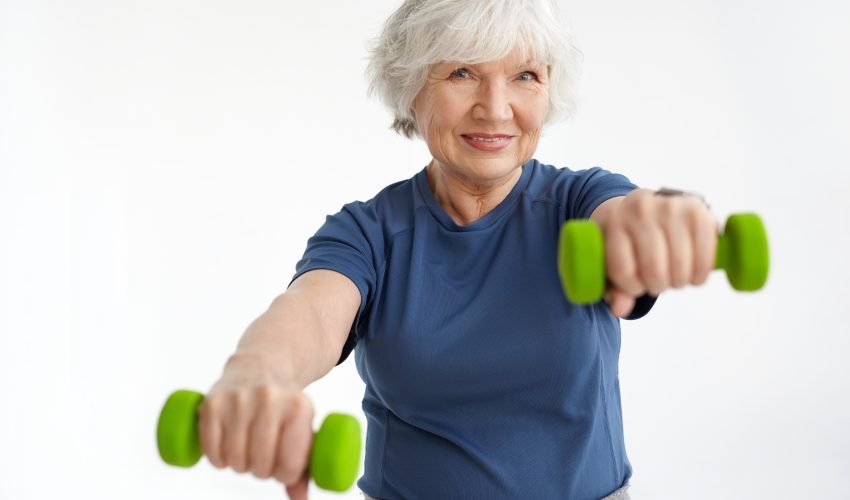 People, age, energy, strength and wellbeing concept. Adorable smiling woman pensioner wearing t-shirt doing physical exercises in the morning, using pair of green dumbbells. Selective focus