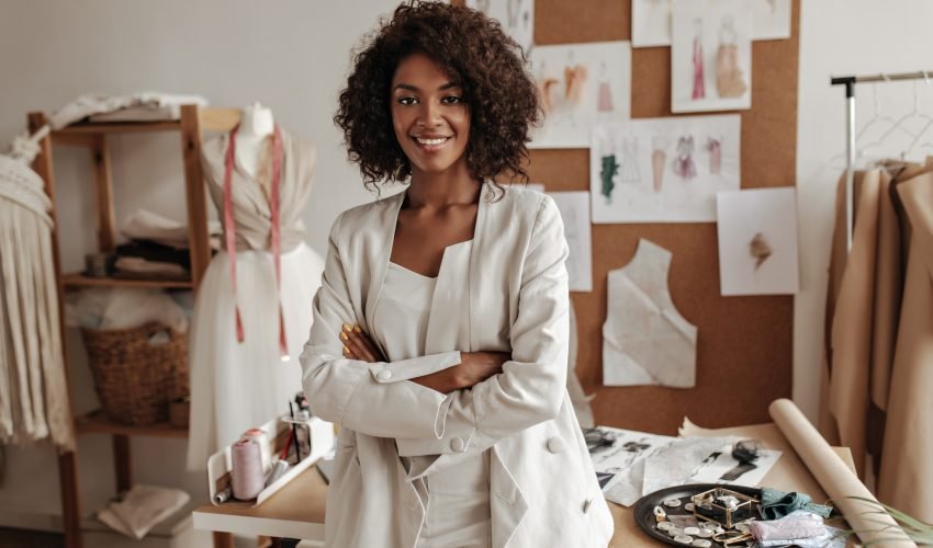 Beautiful curly brunette dark-skinned fashion designer poses in office, leans on table. Young lady