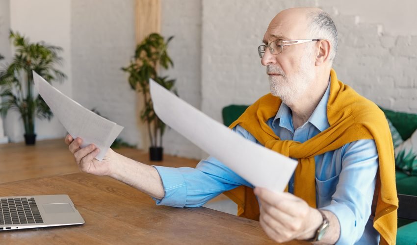 Sad male engineer in his sixties wearing formal clothes and spectacles sitting at wooden desk with generic laptop, holding documents in his hands, being frustrated. Job, occupation and stress concept