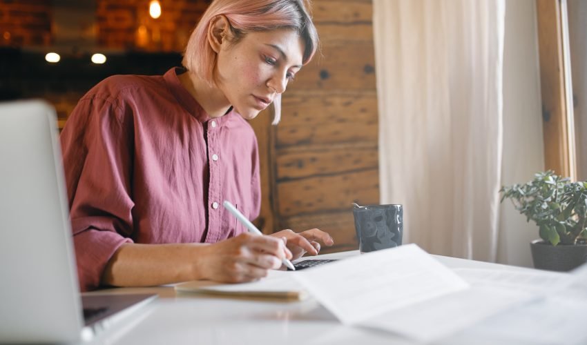 Pensive young office worker checking documentation working from home remotely. Serious woman reading report, sitting at table with laptop. Blonde female calculating expenses, paying bills online