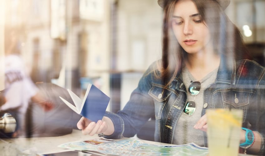 Tourist sitting in cafe reading a map holding passport drinking lemonade in a busy city. Shot through glass.