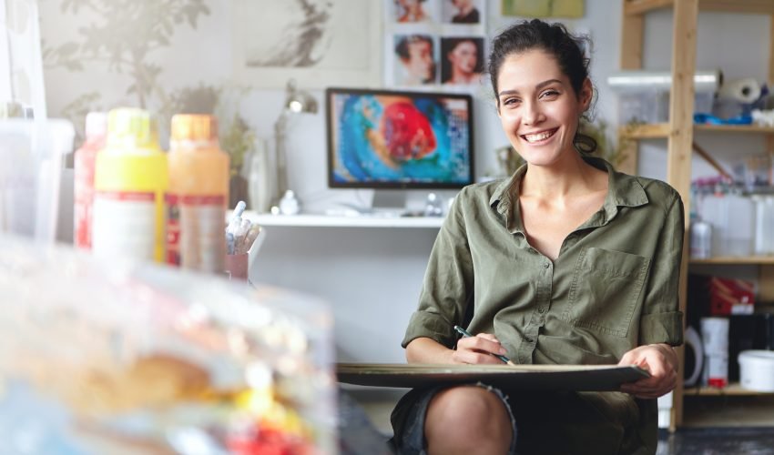Smiling female artist wearing casual clothes sitting in her cabinet with sketches and colorful paint, having happy expression while being glad to create beautiful picture. Painter working in workshop
